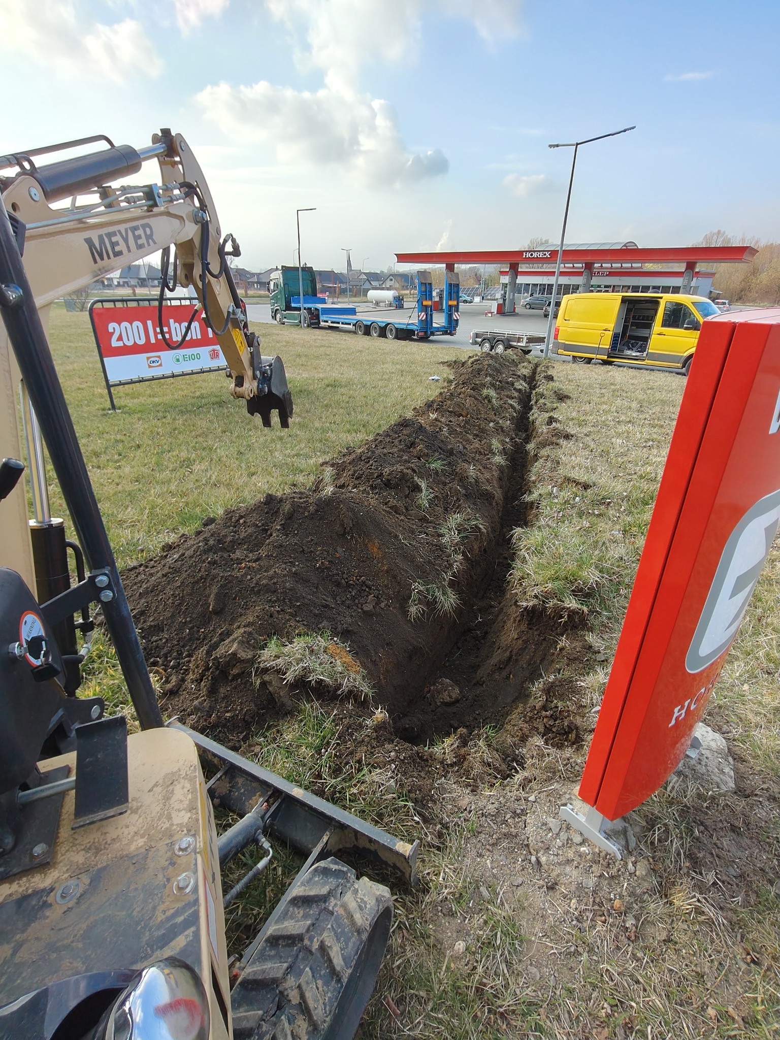 Wykopany rów pod instalację na trawiastym terenie, widoczna koparka Meyer, w tle stacja benzynowa Horex i zaparkowane pojazdy.