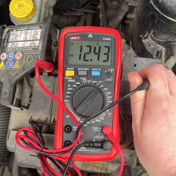 CHECK CAR SPRAWDZENIE SAMOCHODU KRYSTIAN KLIMEK - Pomiar napięcia akumulatora samochodowego za pomocą multimetru UNI-T UT890C, wskazującego 12.43V DC; widoczne przewody pomiarowe i zabrudzone elementy silnika.