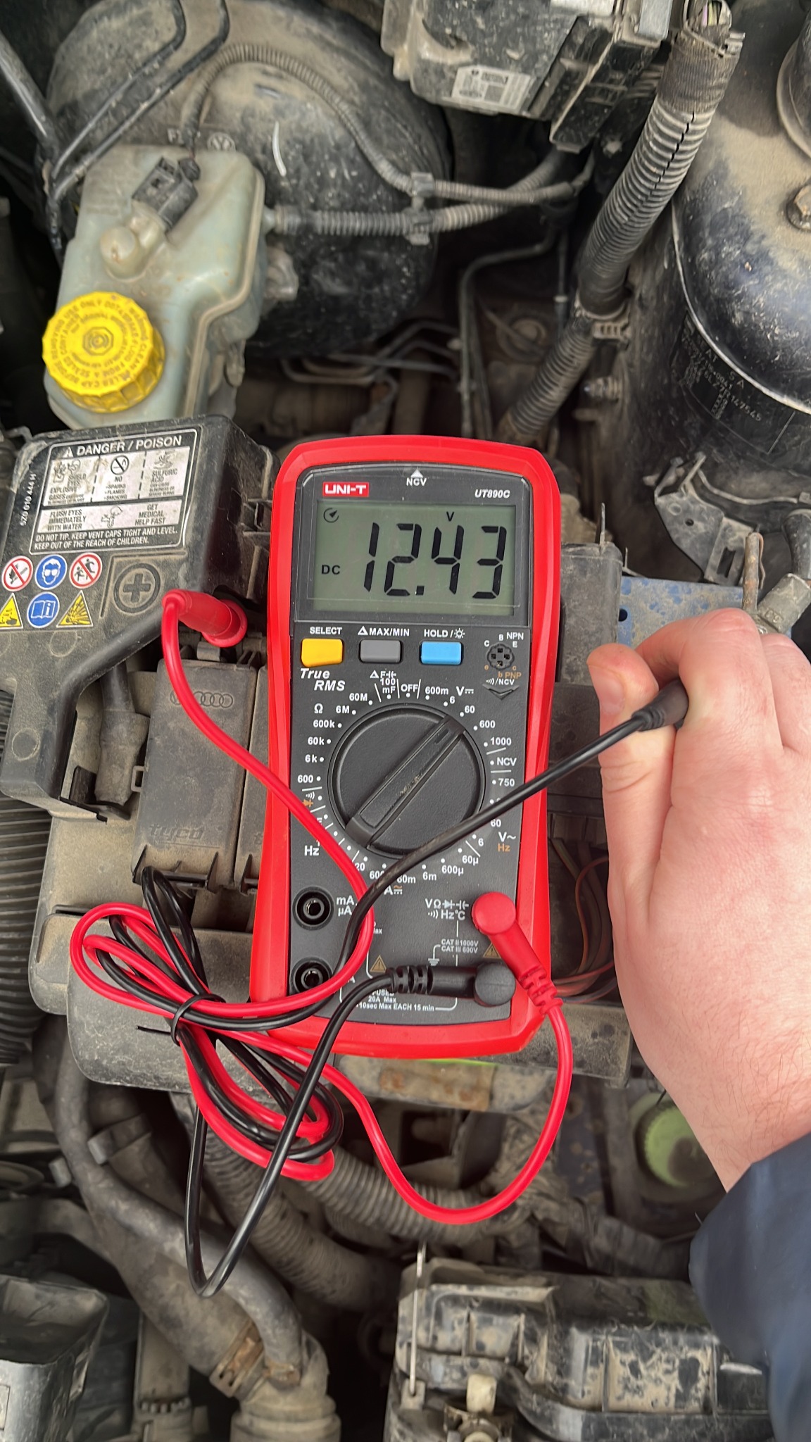 Pomiar napięcia akumulatora samochodowego za pomocą multimetru UNI-T UT890C, wskazującego 12.43V DC; widoczne przewody pomiarowe i zabrudzone elementy silnika.
