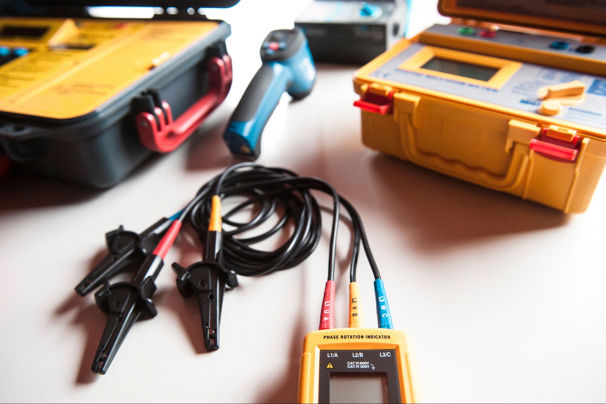 Żółty miernik faz z przewodami zakończonymi krokodylkami, obok skrzynki na narzędzia i termometru bezdotykowego.