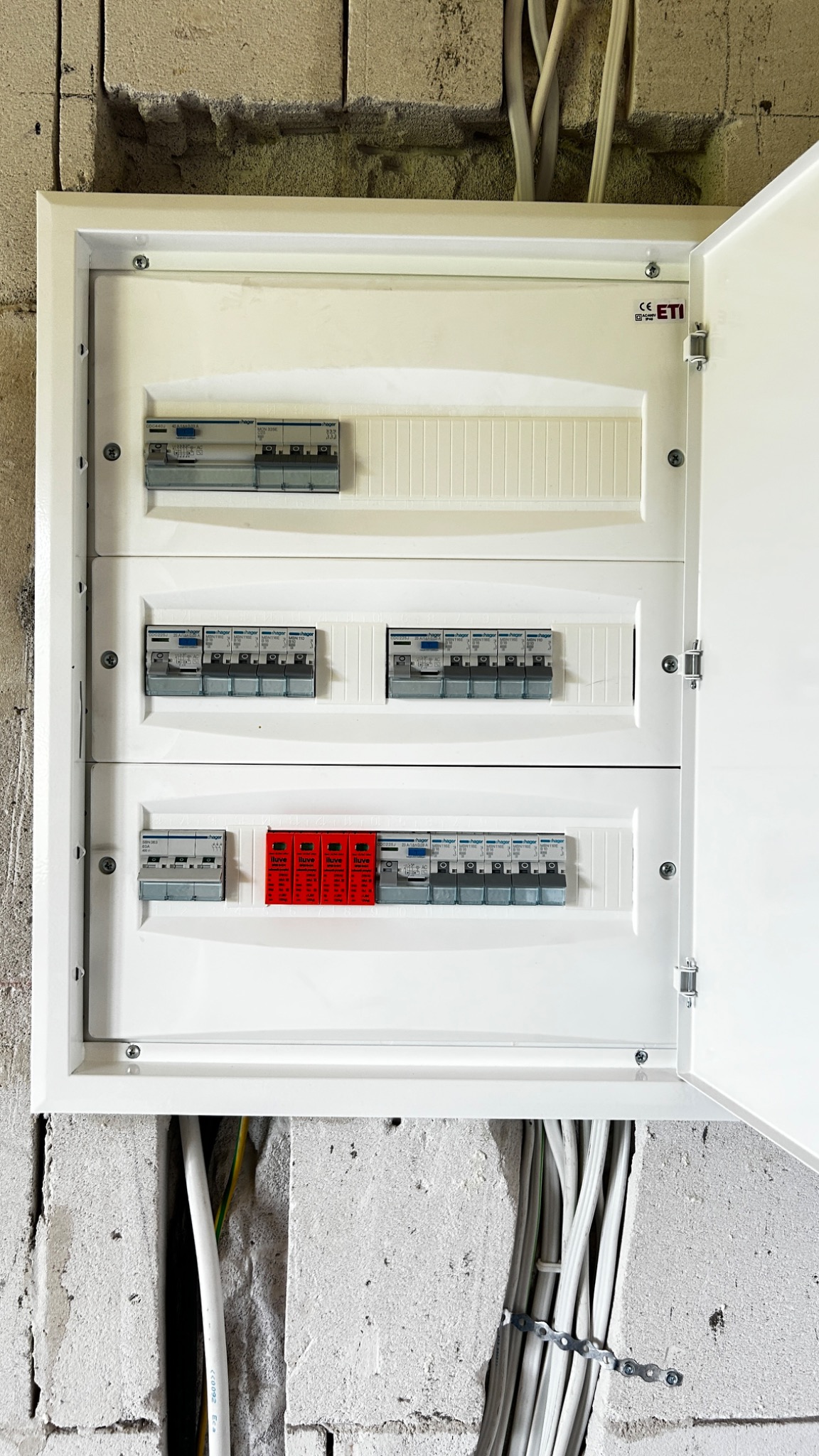 Rozdzielnia elektryczna z otwartymi drzwiczkami, widoczne zabezpieczenia i przewody w surowej ścianie z betonu komórkowego. Instalacja w trakcie realizacji.