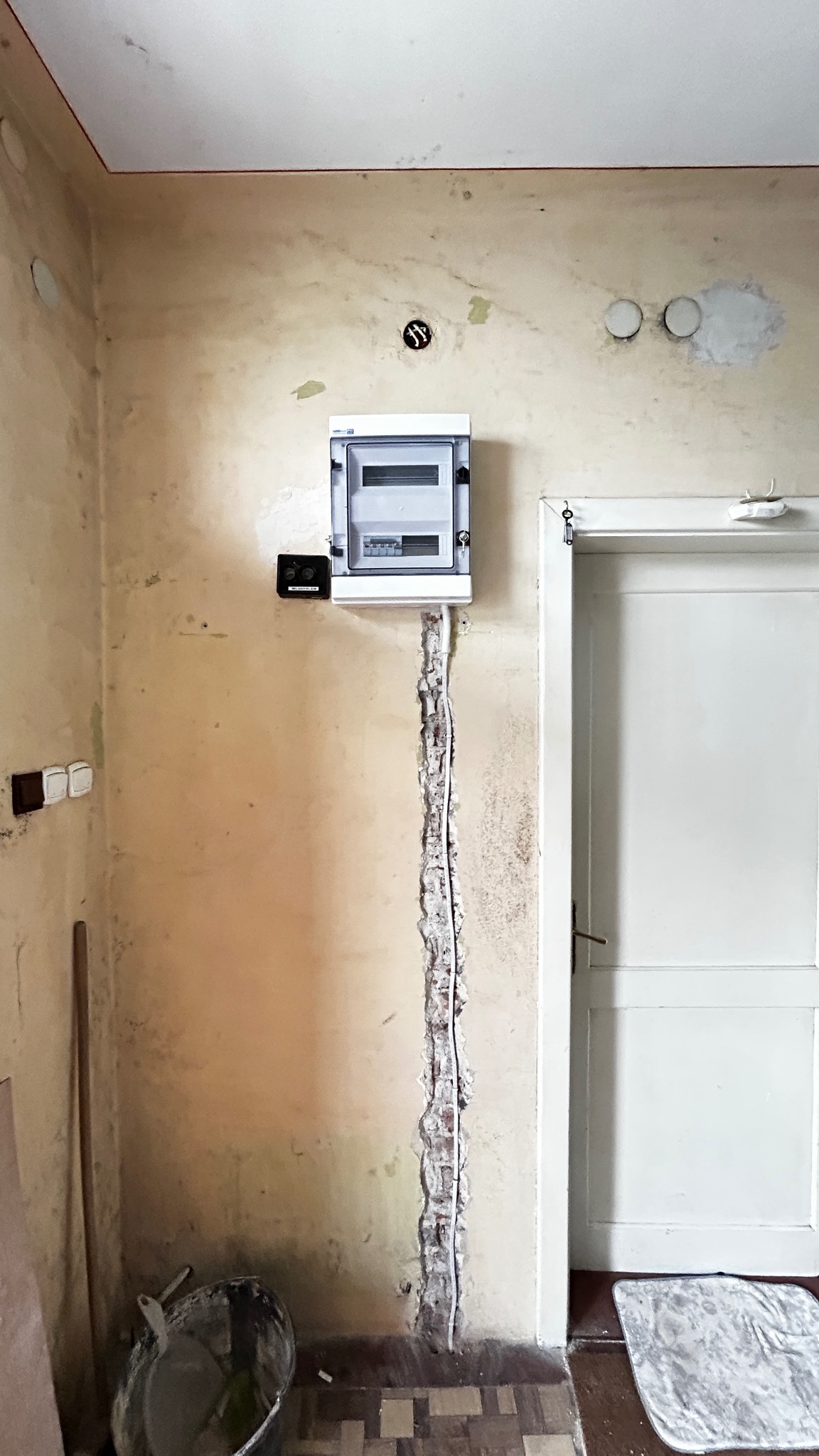 Montaż skrzynki elektrycznej natynkowej w wyżłobieniu ściany, widoczne przewody. Obok białe drzwi i stara instalacja. Ściana w trakcie remontu, widoczne ślady po poprzednich instalacjach.