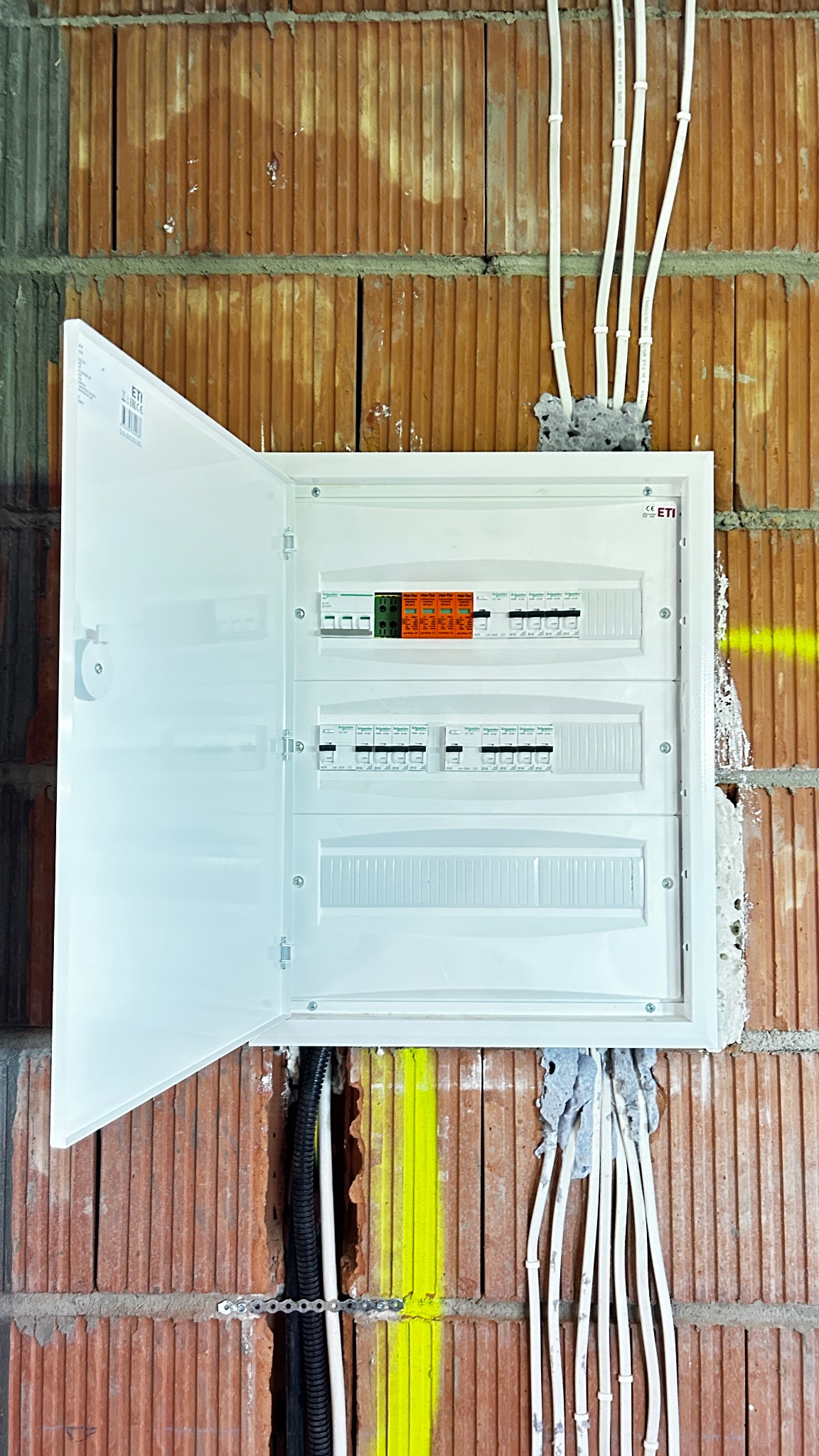 Otwarta, biała skrzynka elektryczna z bezpiecznikami na ceglanej ścianie z wystającymi kablami. Widoczne zabezpieczenia topikowe i wyłączniki nadprądowe.