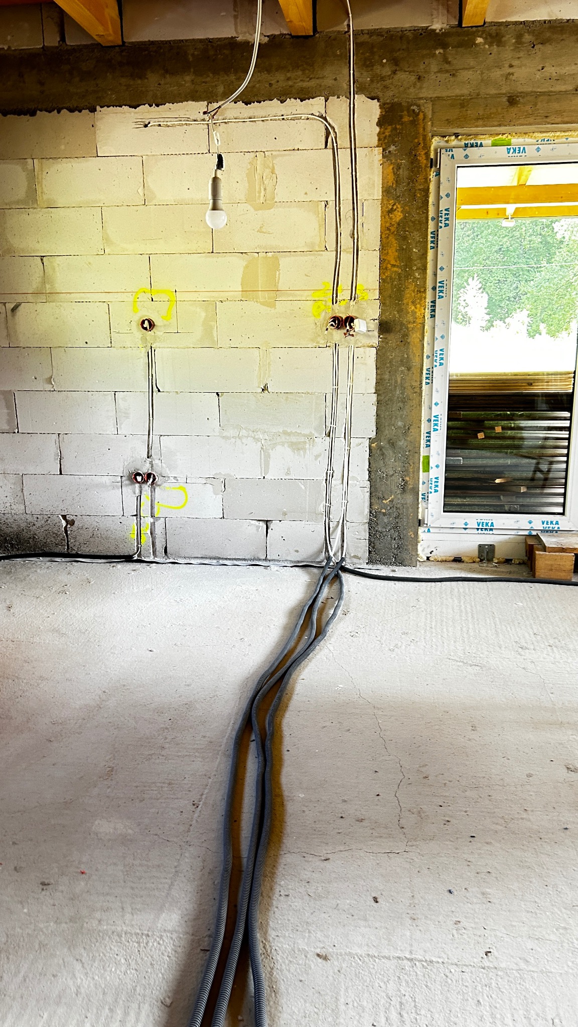 Surowa ściana z betonu komórkowego z przewodami elektrycznymi w peszlach, wystającymi ze ściany. Widoczne puszki instalacyjne. Okno w tle.
