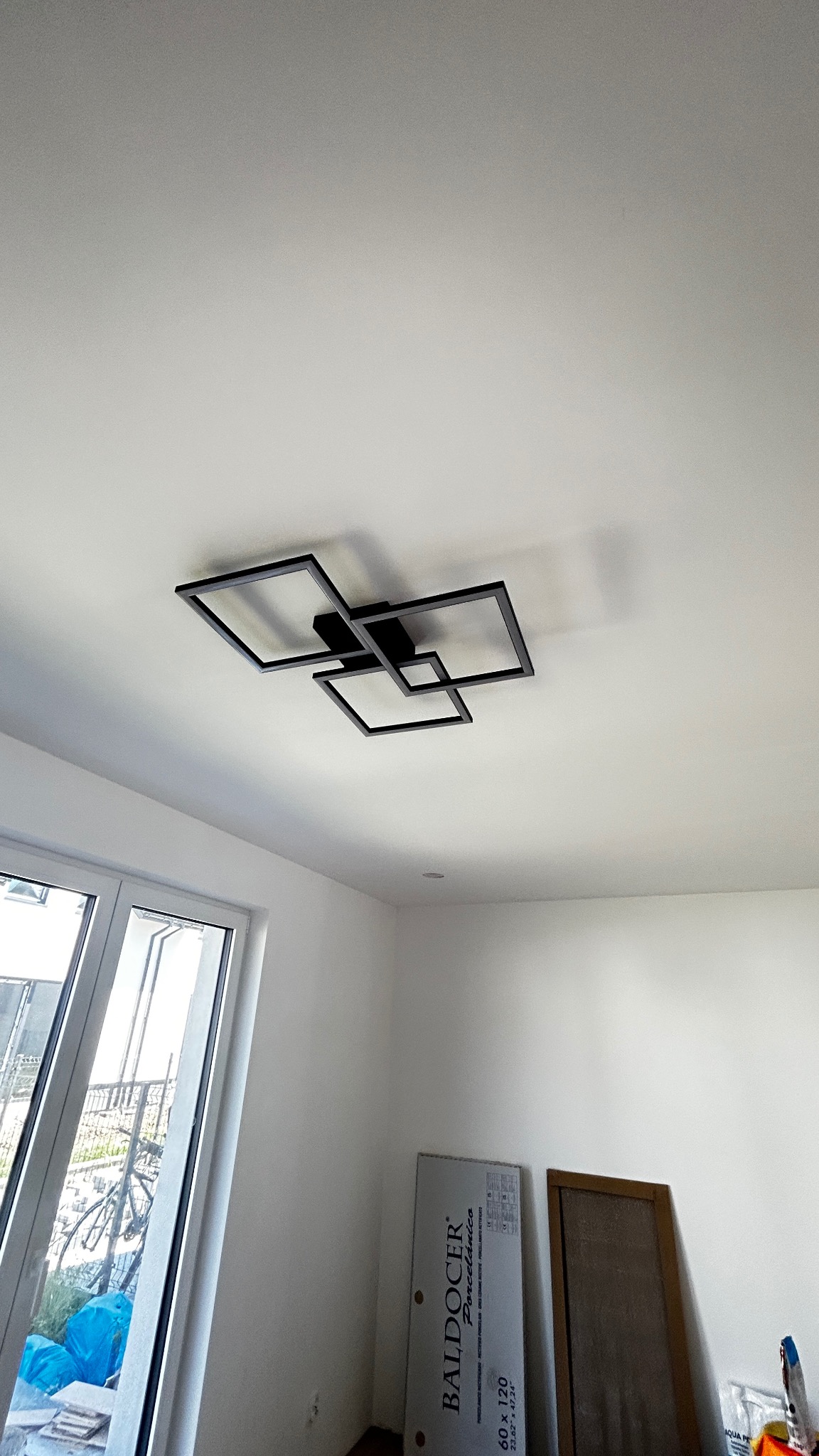 Czarna, geometryczna lampa sufitowa LED w nowoczesnym, minimalistycznym wnętrzu z dużym oknem i elementami płytek podłogowych w tle.