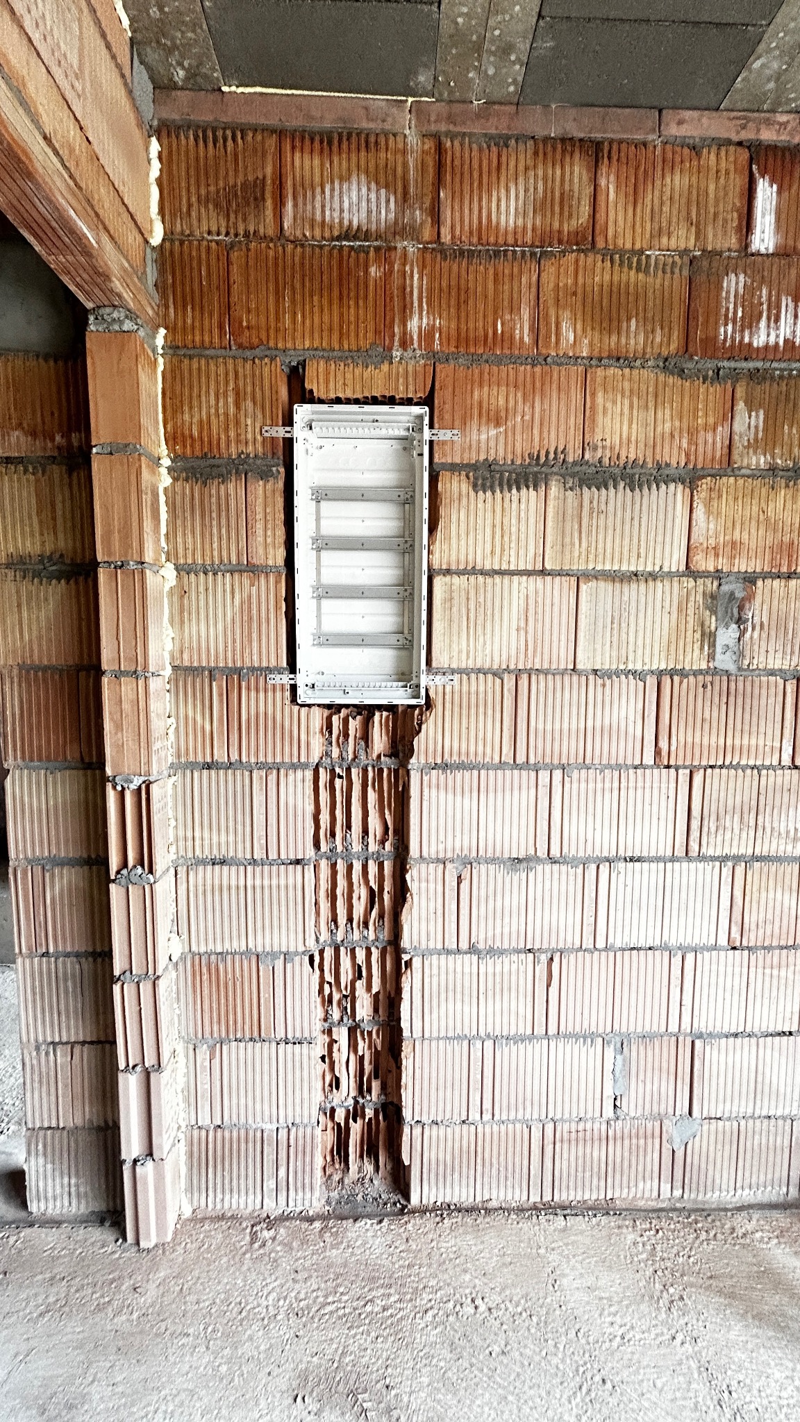 Wnęka w ceglanej ścianie z zamontowaną, białą skrzynką rozdzielczą. Widoczne przewody w przygotowanym kanale. Surowy stan wykończenia.