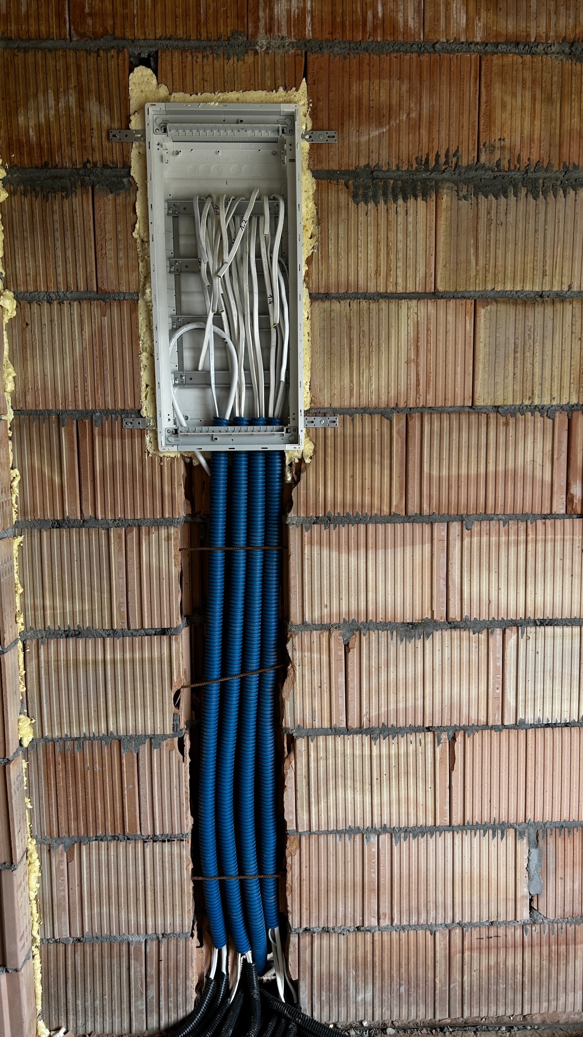 Instalacja elektryczna w trakcie montażu: białe i niebieskie kable prowadzone w ścianie z cegły, zabezpieczone pianką montażową wokół puszki rozdzielczej.
