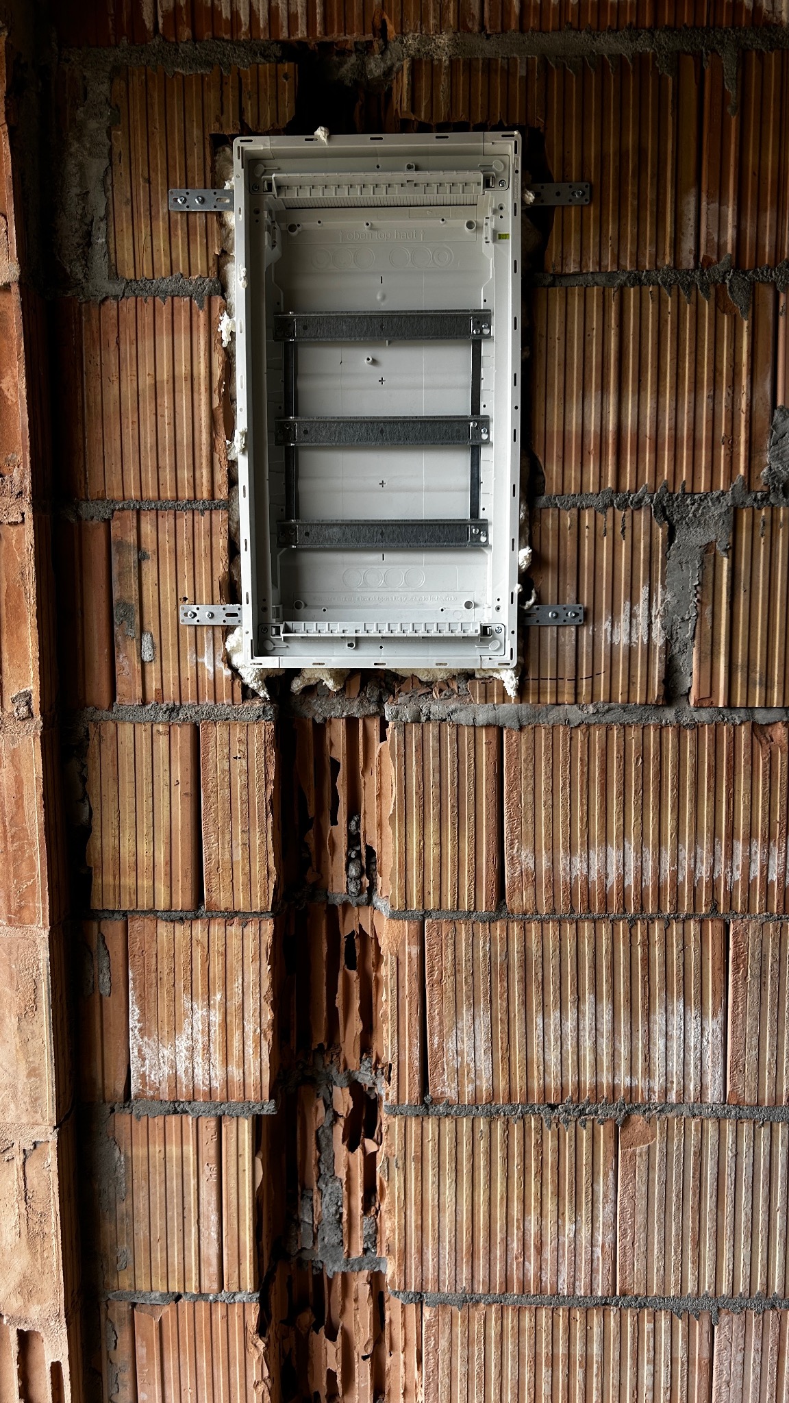 Biała, pusta skrzynka elektryczna zamontowana na ceglanej ścianie z widocznymi śladami wykuwania pod instalację. Widoczne mocowania i pianka montażowa.
