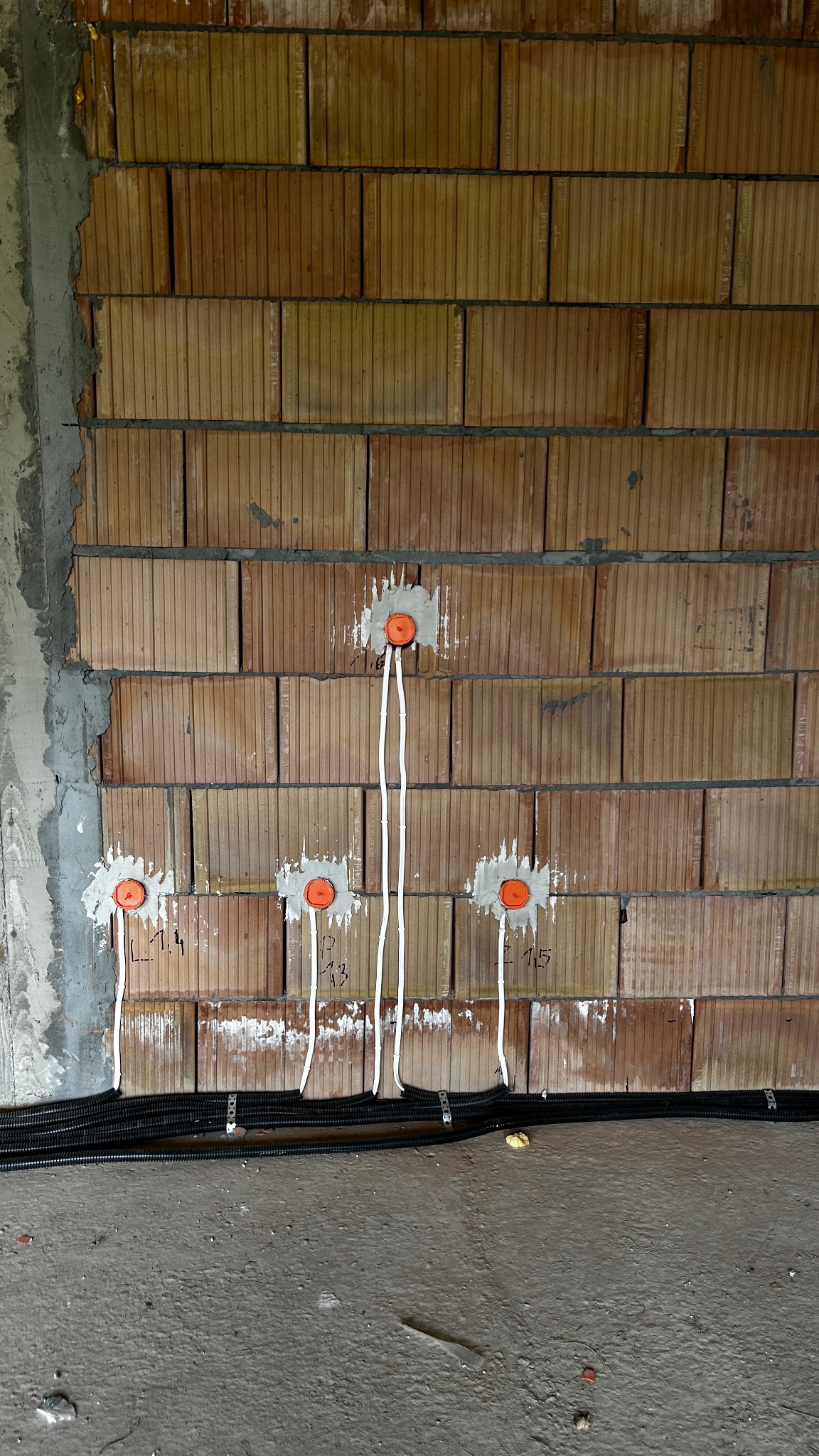 Instalacja elektryczna w trakcie montażu: przewody wychodzące z puszek w surowej, ceglanej ścianie. Widoczne czarne rury instalacyjne przy podłodze.