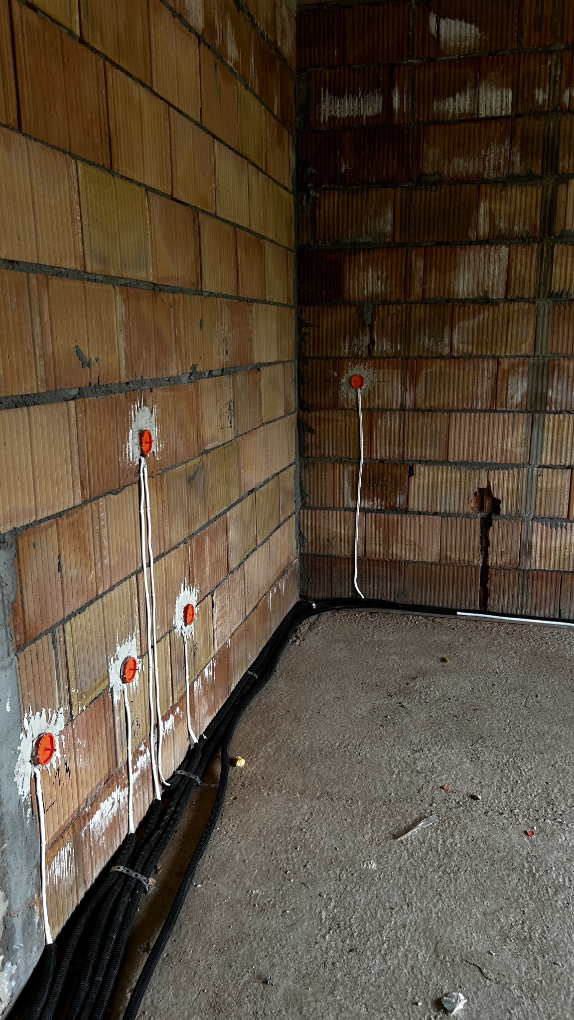 Surowy mur z widocznymi puszkami elektrycznymi i przewodami w peszlach, ułożonymi wzdłuż ściany. Wnętrze w trakcie prac instalacyjnych, widoczne niedoskonałości ścian.
