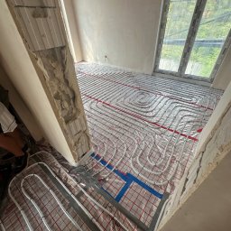 Jakub Szymanik - Układanie rur ogrzewania podłogowego na folii refleksyjnej w surowym pomieszczeniu z oknem dachowym, widoczne fragmenty ściany w stanie deweloperskim z odsłoniętą strukturą.