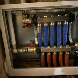 Rozdzielacz instalacji centralnego ogrzewania w metalowej obudowie, z rurami w kolorach niebieskim i czerwonym, widoczne zawory i izolacja na ścianach.