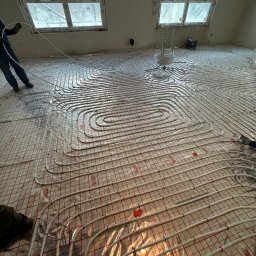 Jakub Szymanik - Układanie rur ogrzewania podłogowego na folii z siatką w nowym budynku z oknami dachowymi, widoczne mocowania rur i napisy 'REFLEX TERMOSYSTEM' na folii.