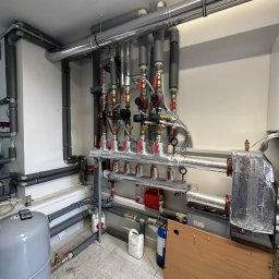 Jakub Szymanik - Rozbudowana instalacja grzewcza w kotłowni: rury stalowe z izolacją, liczne zawory, manometry, naczynie przeponowe, butla z gazem technicznym, widoczne ślady użytkowania na podłodze.