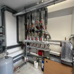 Rozbudowana instalacja grzewcza w kotłowni: rury stalowe z izolacją, liczne zawory, manometry, naczynie przeponowe, butla z gazem technicznym, widoczne ślady użytkowania na podłodze.