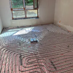 Rozłożona instalacja ogrzewania podłogowego z rur na folii z czerwoną siatką w surowym wnętrzu, widoczne okno z widokiem na zieleń.