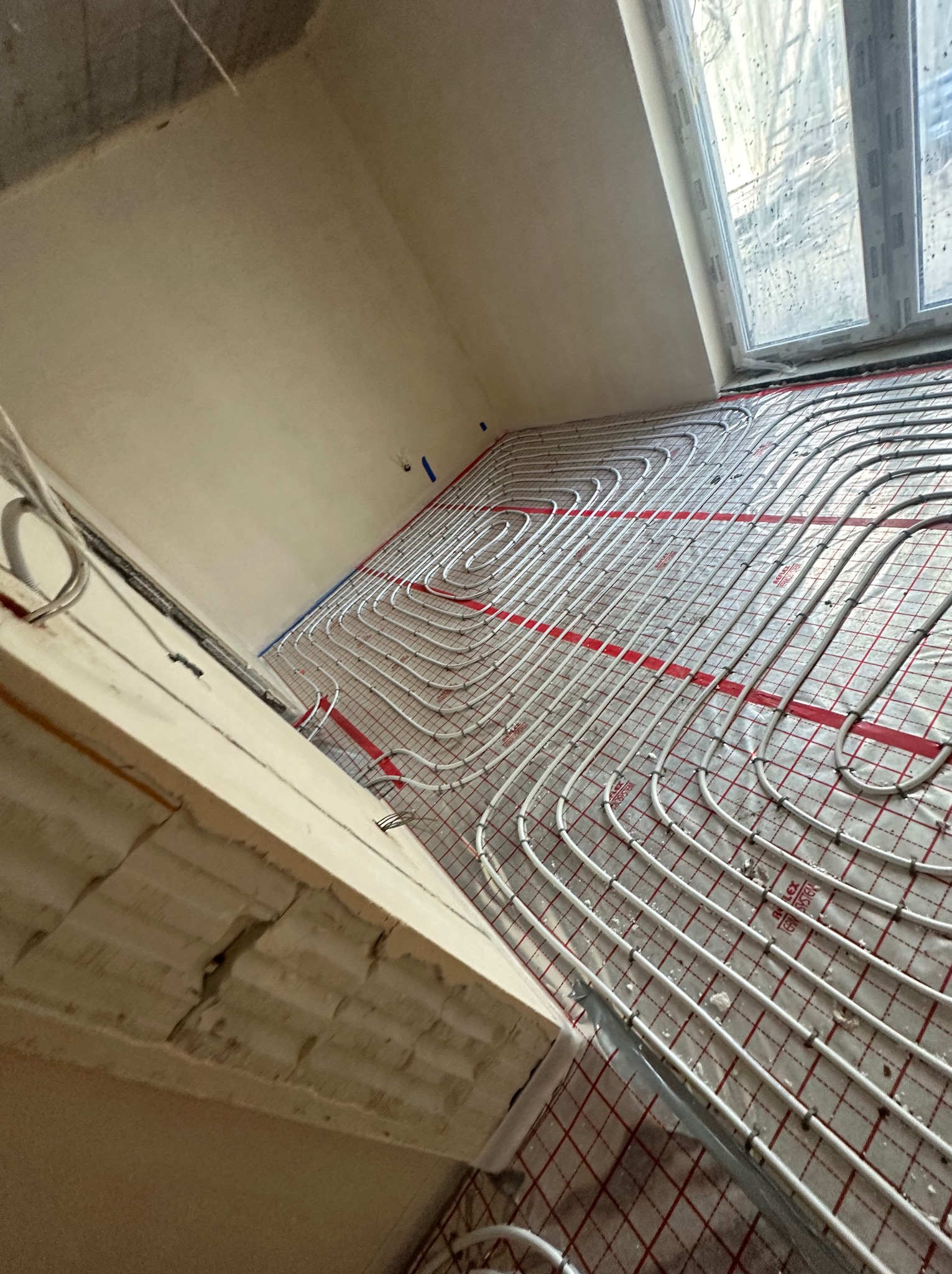Rozłożone rury ogrzewania podłogowego na czerwonej siatce w surowym wnętrzu, widoczne okno i ściany w trakcie prac wykończeniowych.