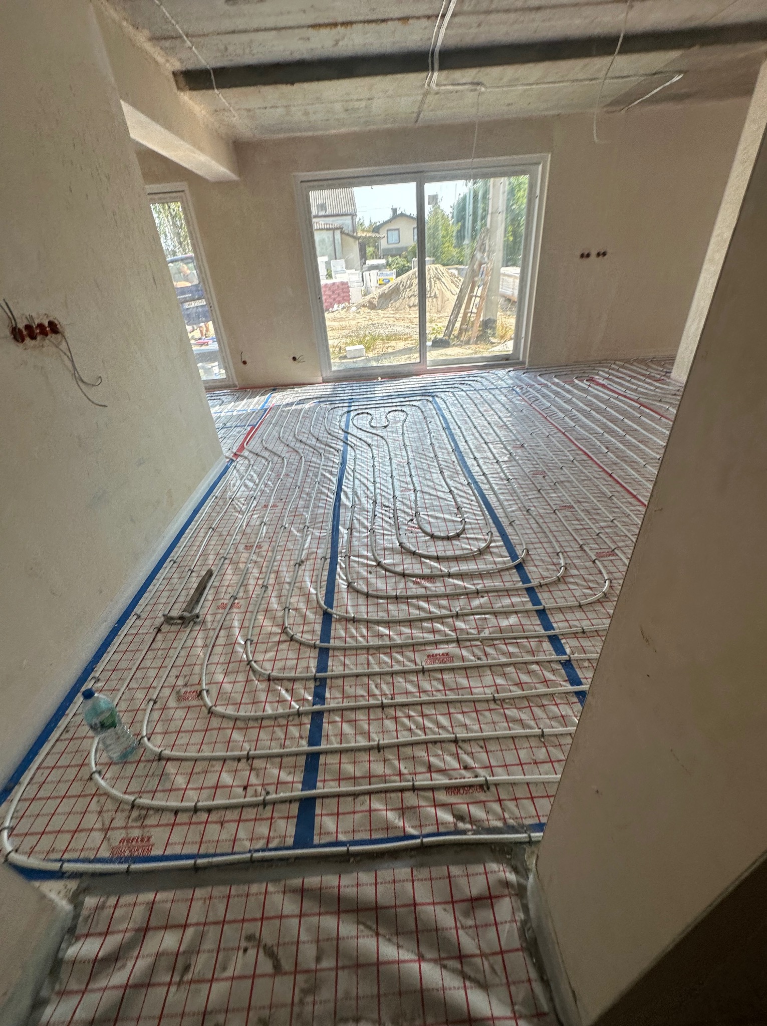 Rozłożona instalacja ogrzewania podłogowego w surowym wnętrzu, widoczne białe rury na czerwonej macie z niebieskimi taśmami, butelka wody i kombinerki przy ścianie, okno z widokiem na budowę.