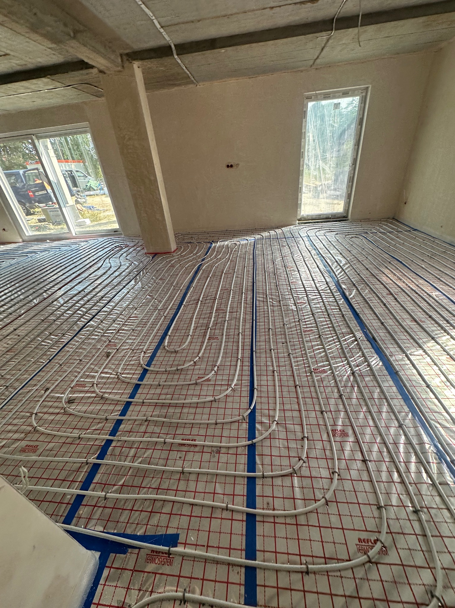 Rozłożone białe rurki ogrzewania podłogowego na folii z czerwoną siatką w surowym stanie budynku, widoczne okna i betonowy słup.