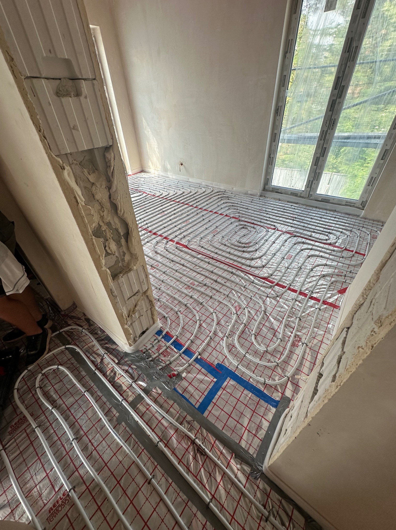 Układanie rur ogrzewania podłogowego na folii refleksyjnej w surowym pomieszczeniu z oknem dachowym, widoczne fragmenty ściany w stanie deweloperskim z odsłoniętą strukturą.