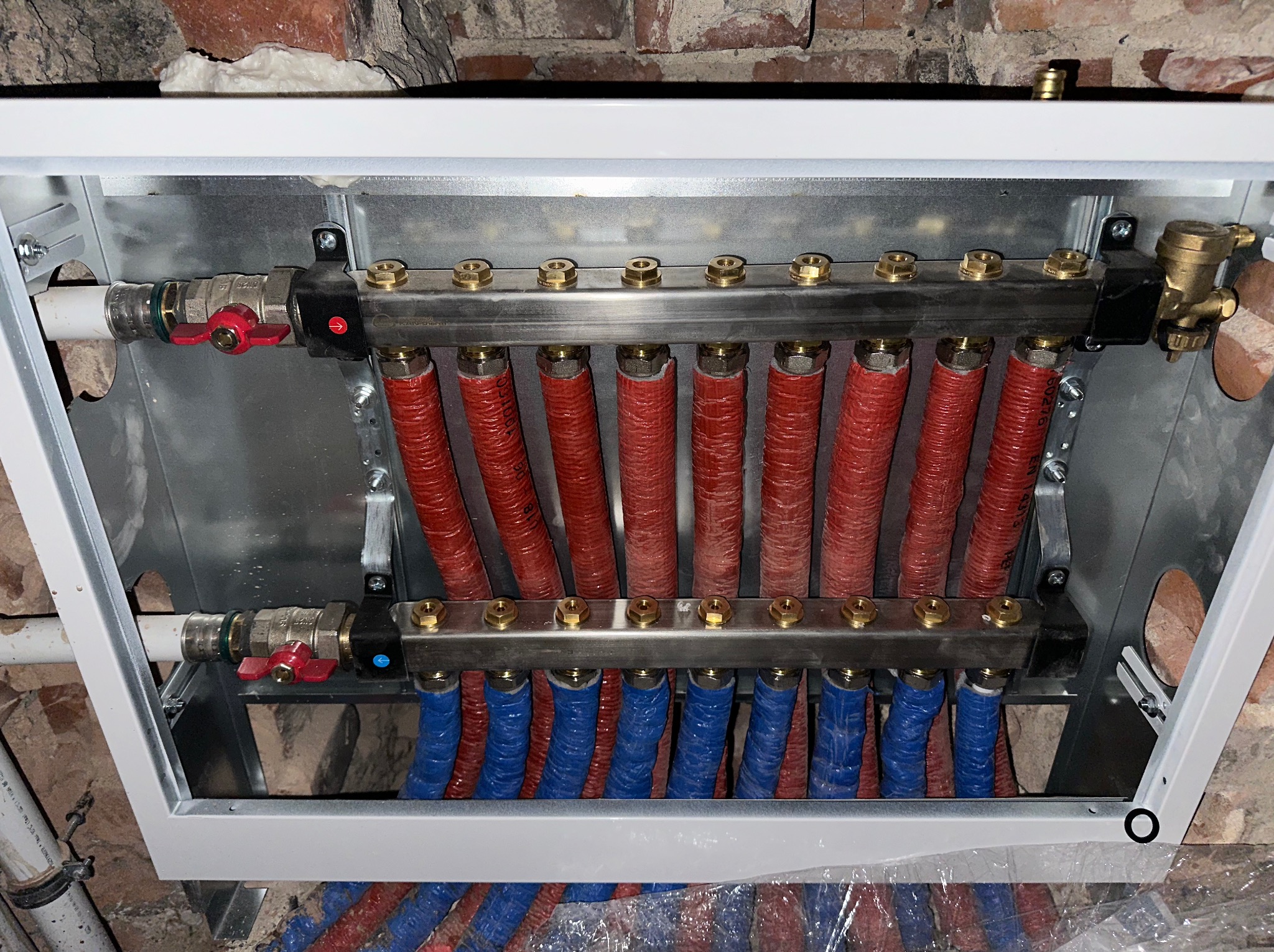 Rozdzielacz instalacji grzewczej podłączony do rur w kolorach czerwonym i niebieskim, zamontowany w metalowej obudowie na tle ceglanej ściany.