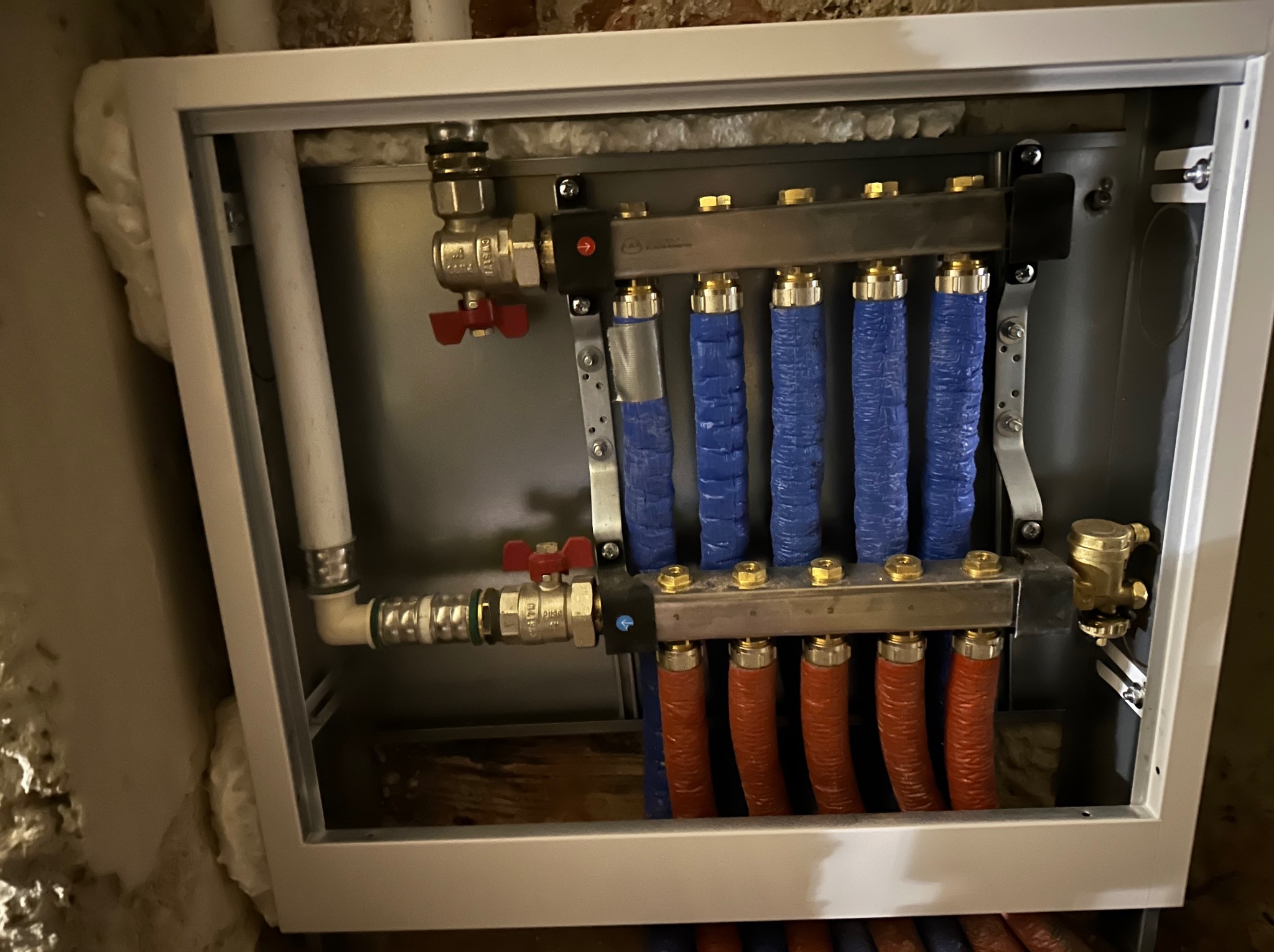 Rozdzielacz instalacji centralnego ogrzewania w metalowej obudowie, z rurami w kolorach niebieskim i czerwonym, widoczne zawory i izolacja na ścianach.