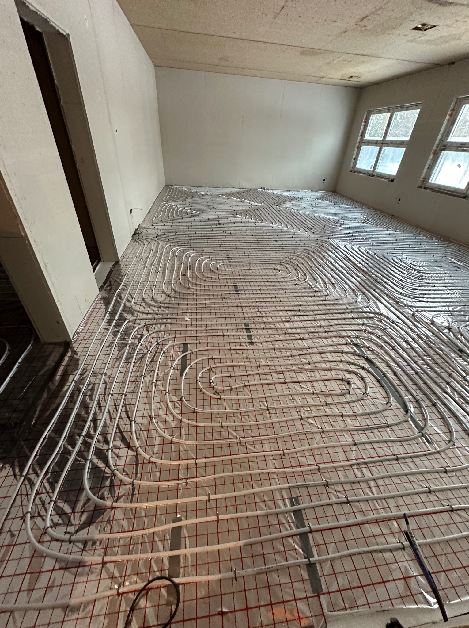 Białe rury ogrzewania podłogowego ułożone spiralnie na folii z czerwoną siatką w surowym pomieszczeniu z oknami.