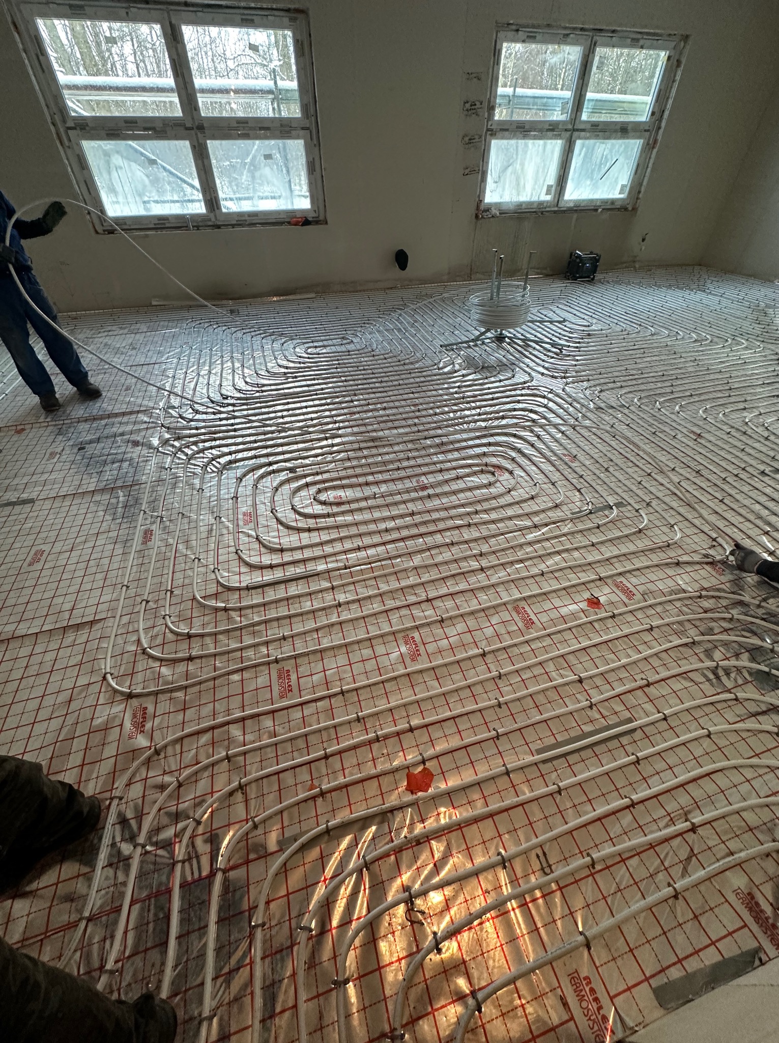 Układanie rur ogrzewania podłogowego na folii z siatką w nowym budynku z oknami dachowymi, widoczne mocowania rur i napisy 'REFLEX TERMOSYSTEM' na folii.
