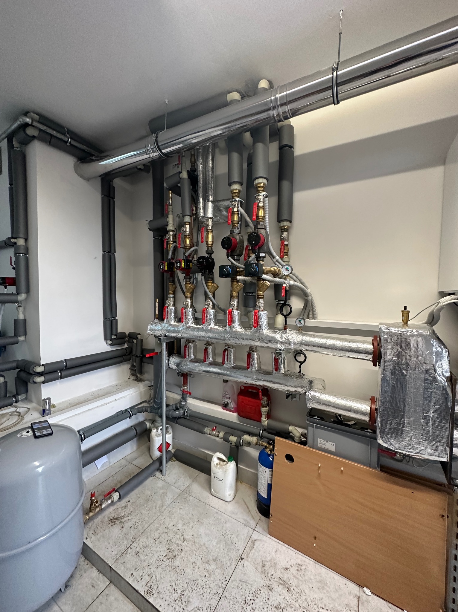 Rozbudowana instalacja grzewcza w kotłowni: rury stalowe z izolacją, liczne zawory, manometry, naczynie przeponowe, butla z gazem technicznym, widoczne ślady użytkowania na podłodze.