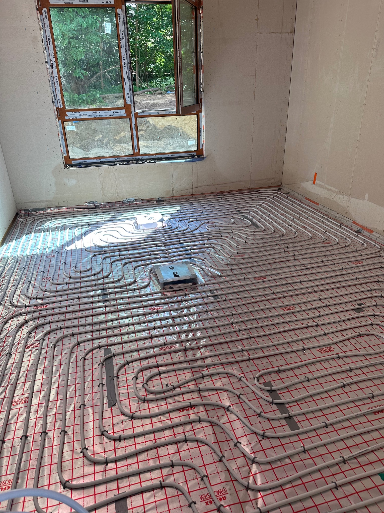 Rozłożona instalacja ogrzewania podłogowego z rur na folii z czerwoną siatką w surowym wnętrzu, widoczne okno z widokiem na zieleń.