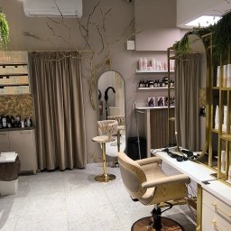 Biały kowal - Wnętrze nowoczesnego salonu fryzjerskiego z beżowymi fotelami, złotymi akcentami, lustrami i dekoracyjnym drzewem na ścianie. Widoczne oświetlenie punktowe i jasna podłoga z terazzo.