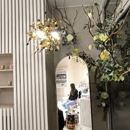 Biały kowal - Odbicie w lustrze salonu kosmetycznego z klientką siedzącą na krześle podczas zabiegu manicure, widoczne dekoracje kwiatowe i lampa wisząca.