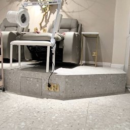 Biały kowal - Stanowisko do pedicure z szarym fotelem, białą lampą i stolikiem, podest wykończony płytkami lastryko, dekoracja z gałęzi z kwiatami.