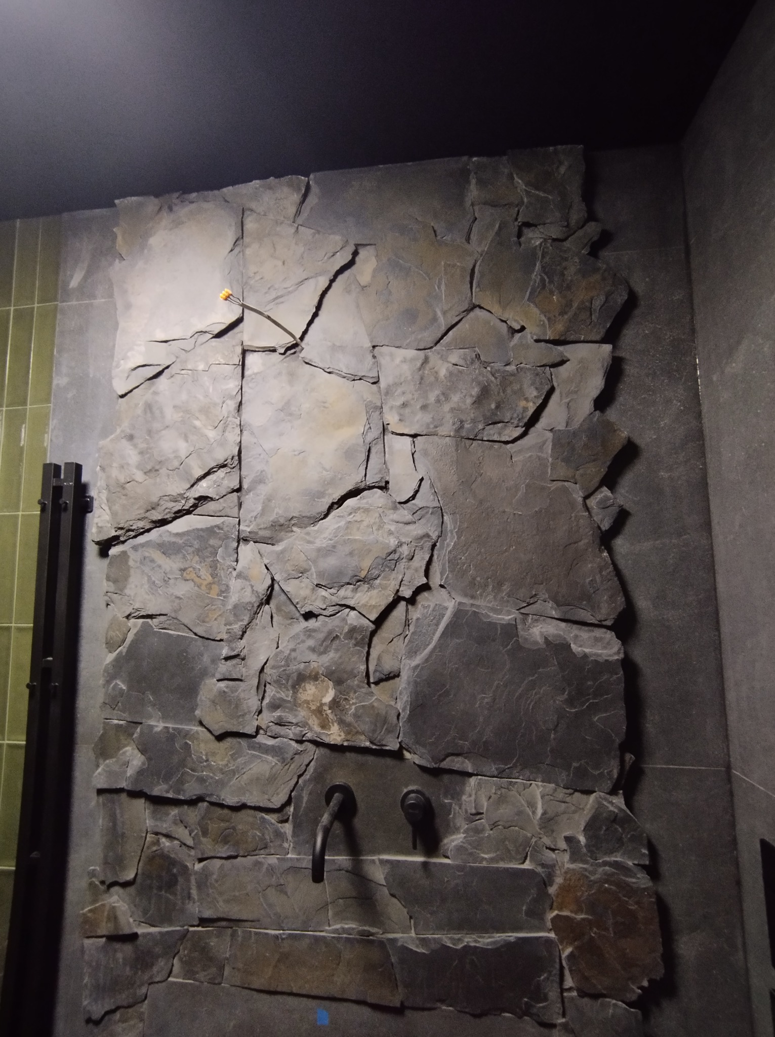 Ściana w łazience wykończona szarym kamieniem łupkowym o nieregularnych kształtach, z widoczną czarną armaturą w stylu retro i fragmentem zielonej ściany z płytek w tle.