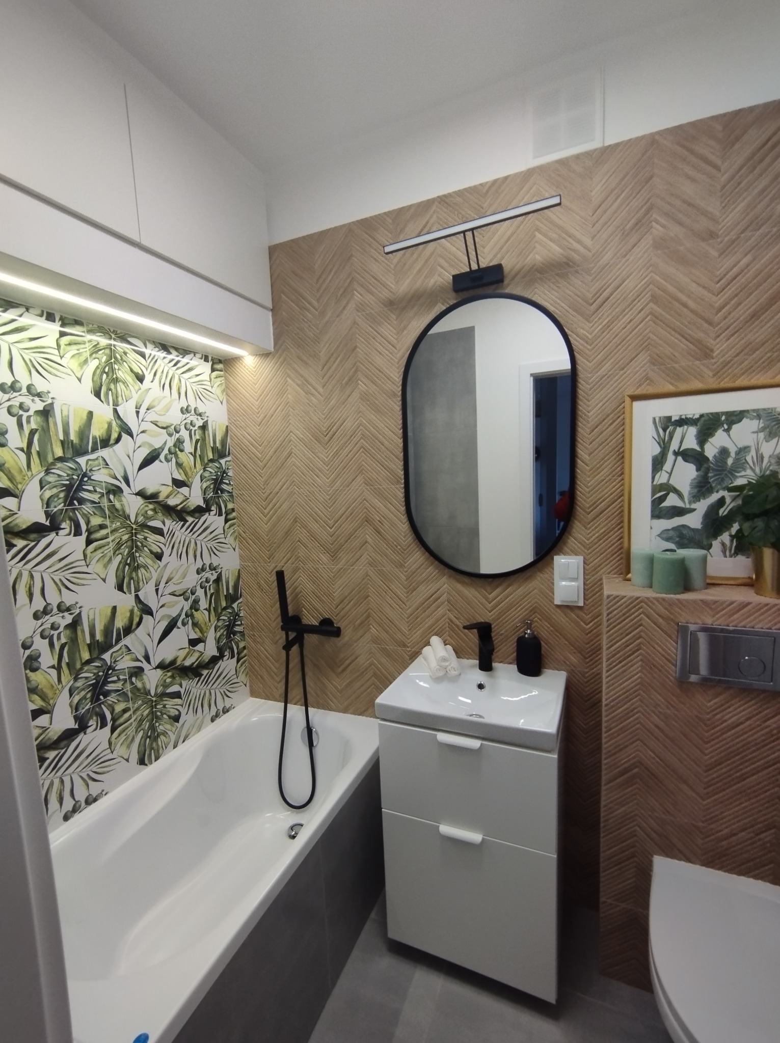 Nowoczesna łazienka z wanną obłożoną płytkami w tropikalne liście, umywalką na szafce z czarną armaturą i lustrem owalnym, oraz płytkami imitującymi drewno ułożonymi w jodełkę na ścianie.