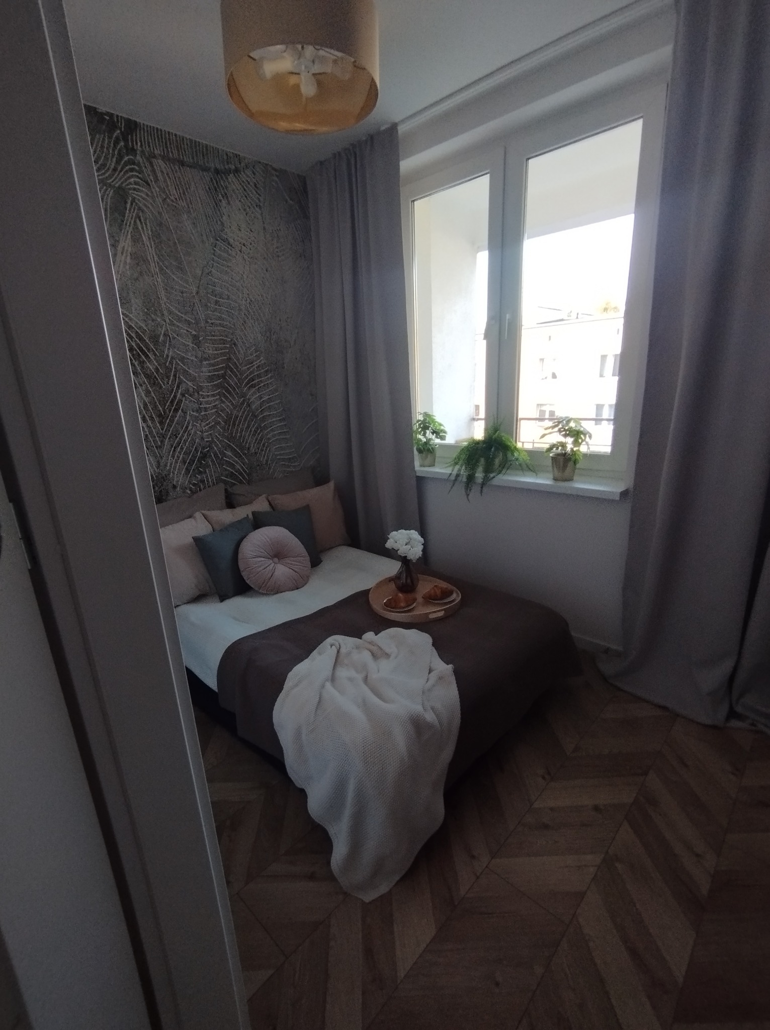 Przytulna sypialnia z parkietem w jodełkę, łóżkiem z dekoracyjną poduszką i tacą z przekąskami, fototapeta z motywem roślinnym i rośliny doniczkowe na parapecie okna.