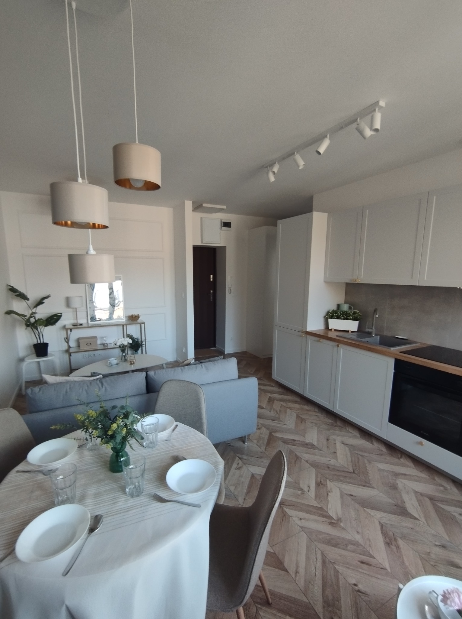Jasne wnętrze mieszkania z parkietem ułożonym w jodełkę, widok na salon z szarą sofą, okrągły stół z nakryciem i kuchnię z białymi szafkami.
