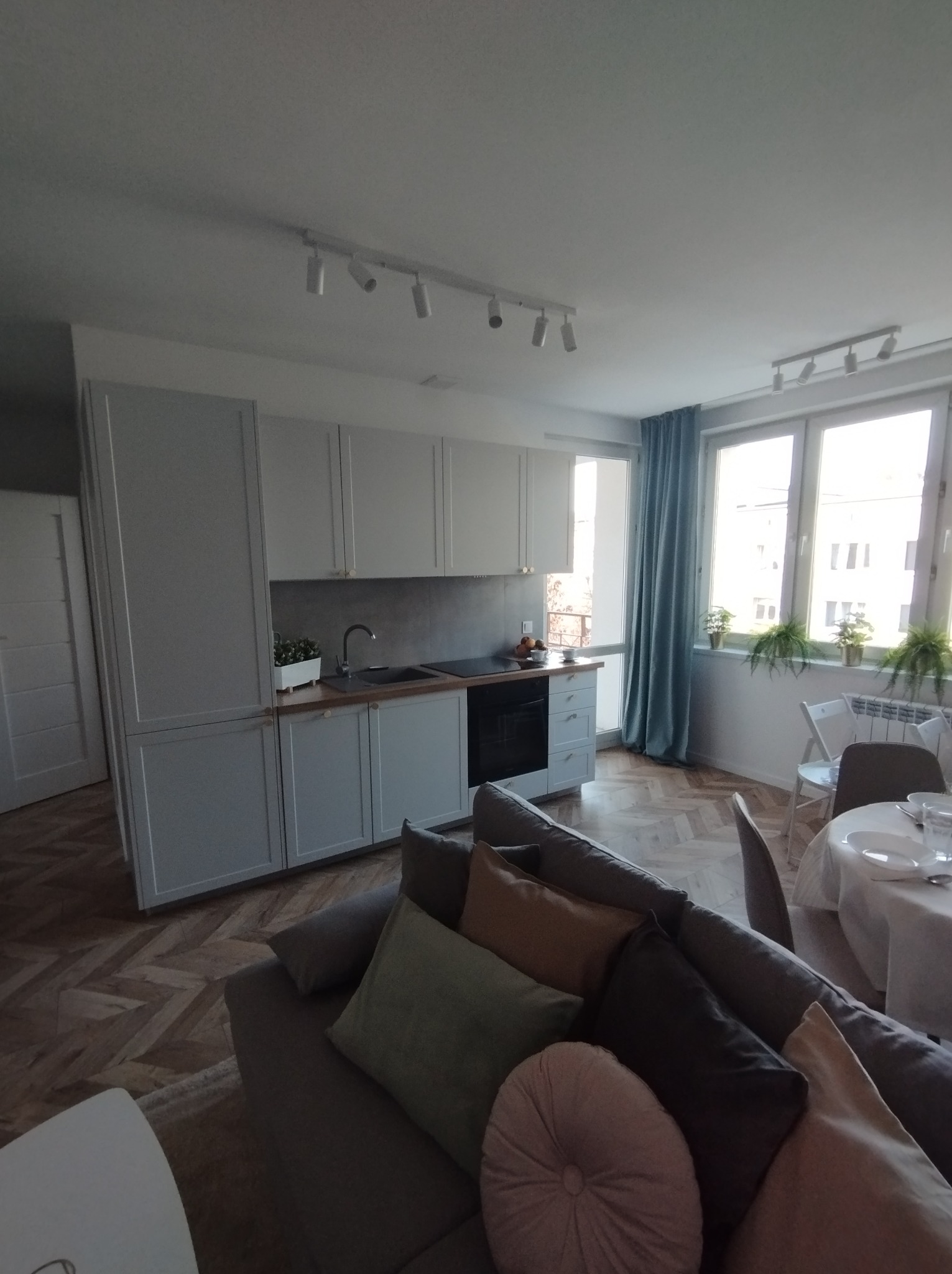 Jasna kuchnia z szarymi szafkami i drewnianym blatem, widok z salonu z szarą sofą i okrągłym stołem nakrytym do posiłku, parkiet ułożony w jodełkę.