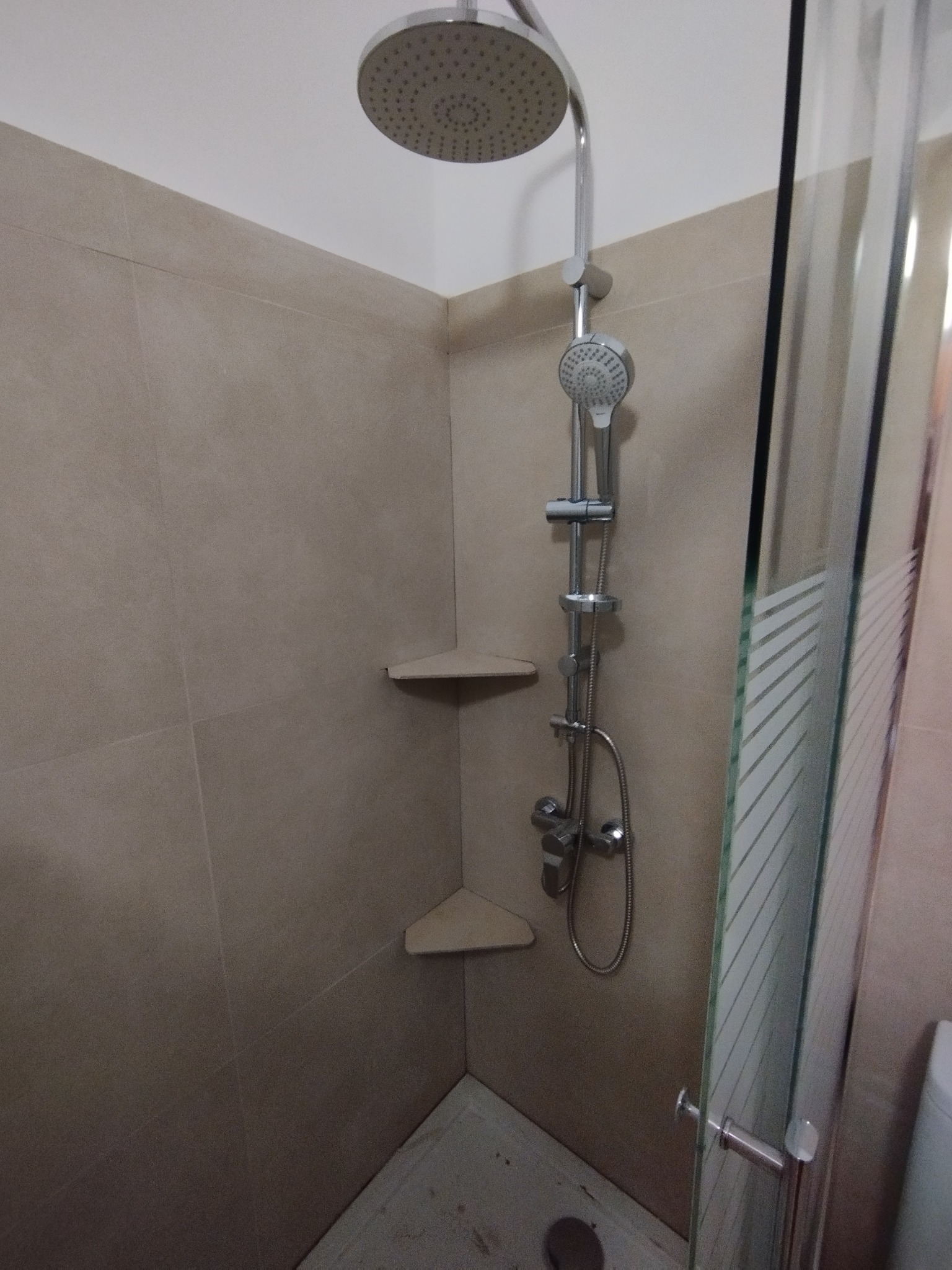 Wykończona kabina prysznicowa z deszczownicą i półkami narożnymi, zamknięta szklanymi drzwiami z poziomymi paskami, wykończona beżowymi płytkami.