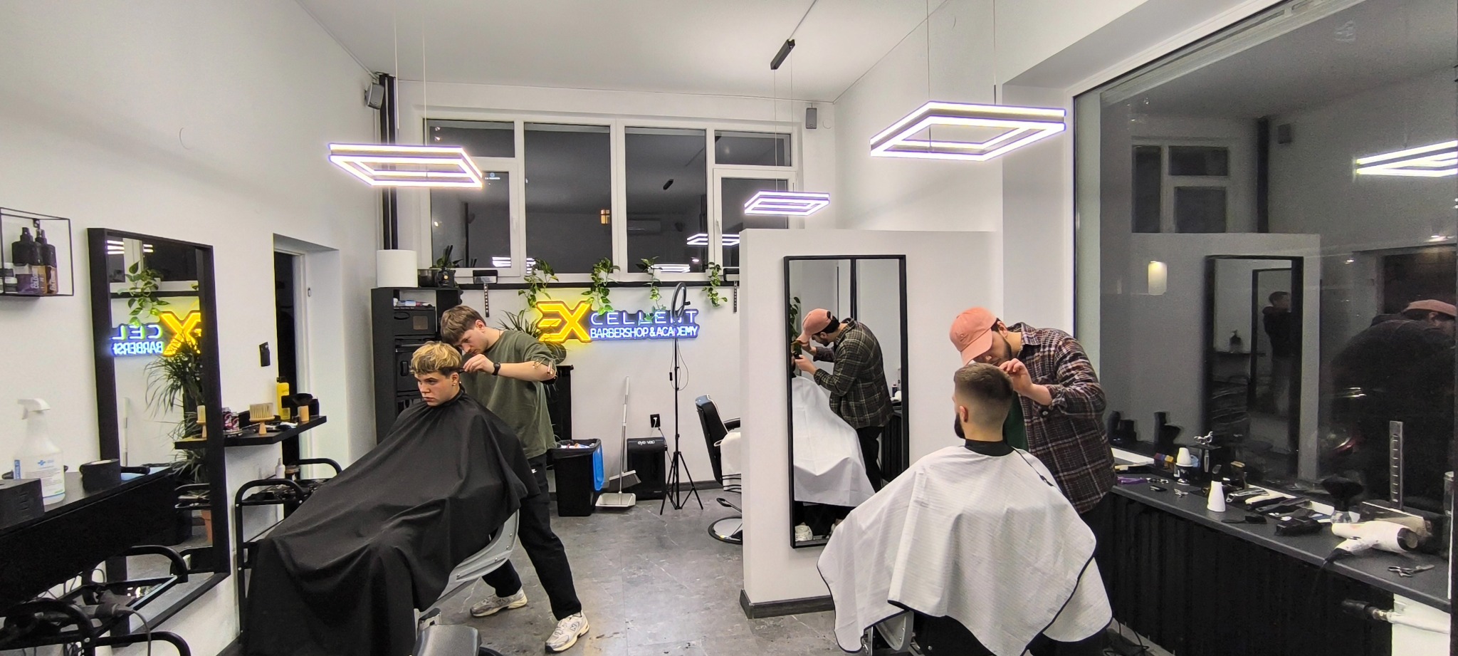 Wnętrze nowoczesnego salonu fryzjerskiego z dwoma fryzjerami strzyżącymi klientów; widoczne lustra, oświetlenie LED i logo salonu na ścianie.