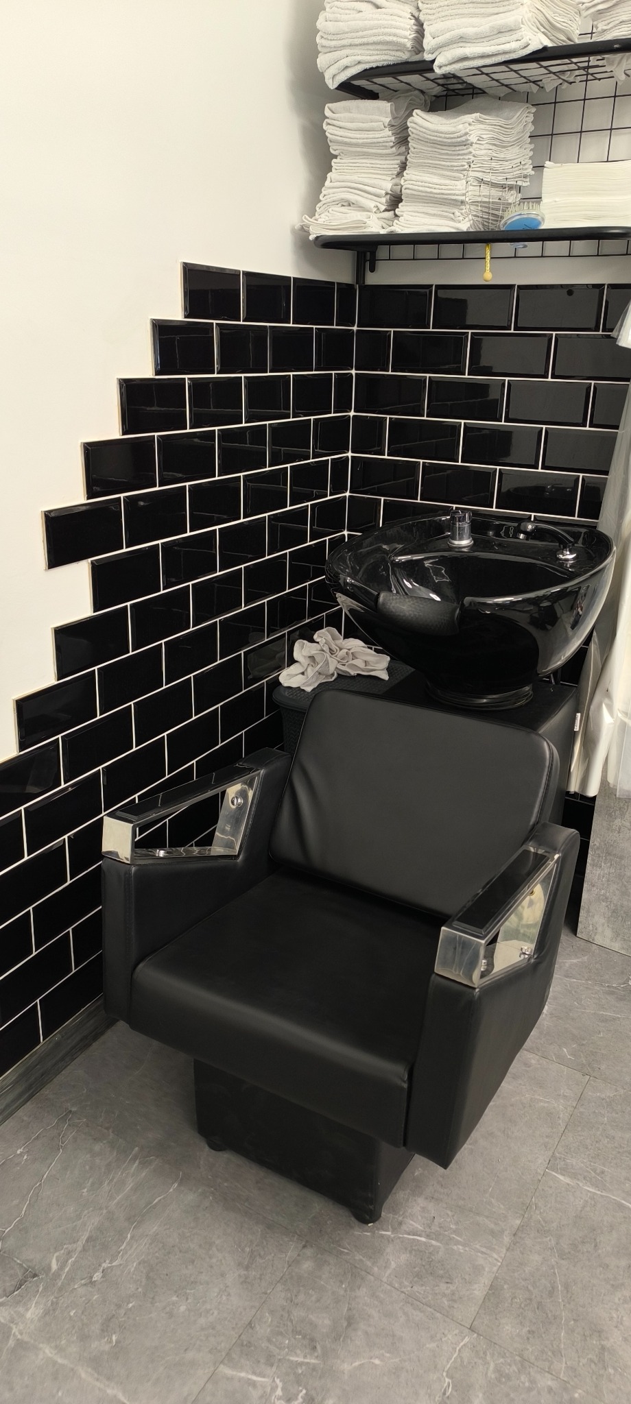 Czarne płytki ułożone schodkowo na ścianie w łazience, z czarną umywalką i fotelem fryzjerskim z chromowanymi podłokietnikami, obok stos ręczników.