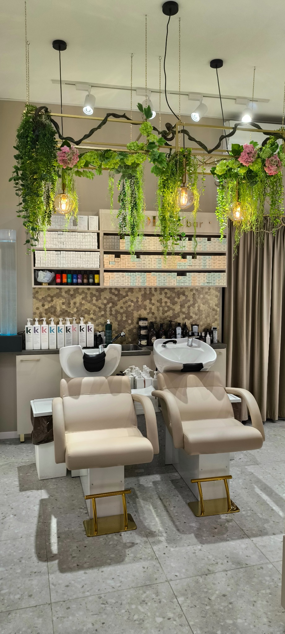 Dwa stanowiska do mycia włosów z beżowymi fotelami w salonie fryzjerskim, z dekoracjami roślinnymi i oświetleniem w stylu loft.