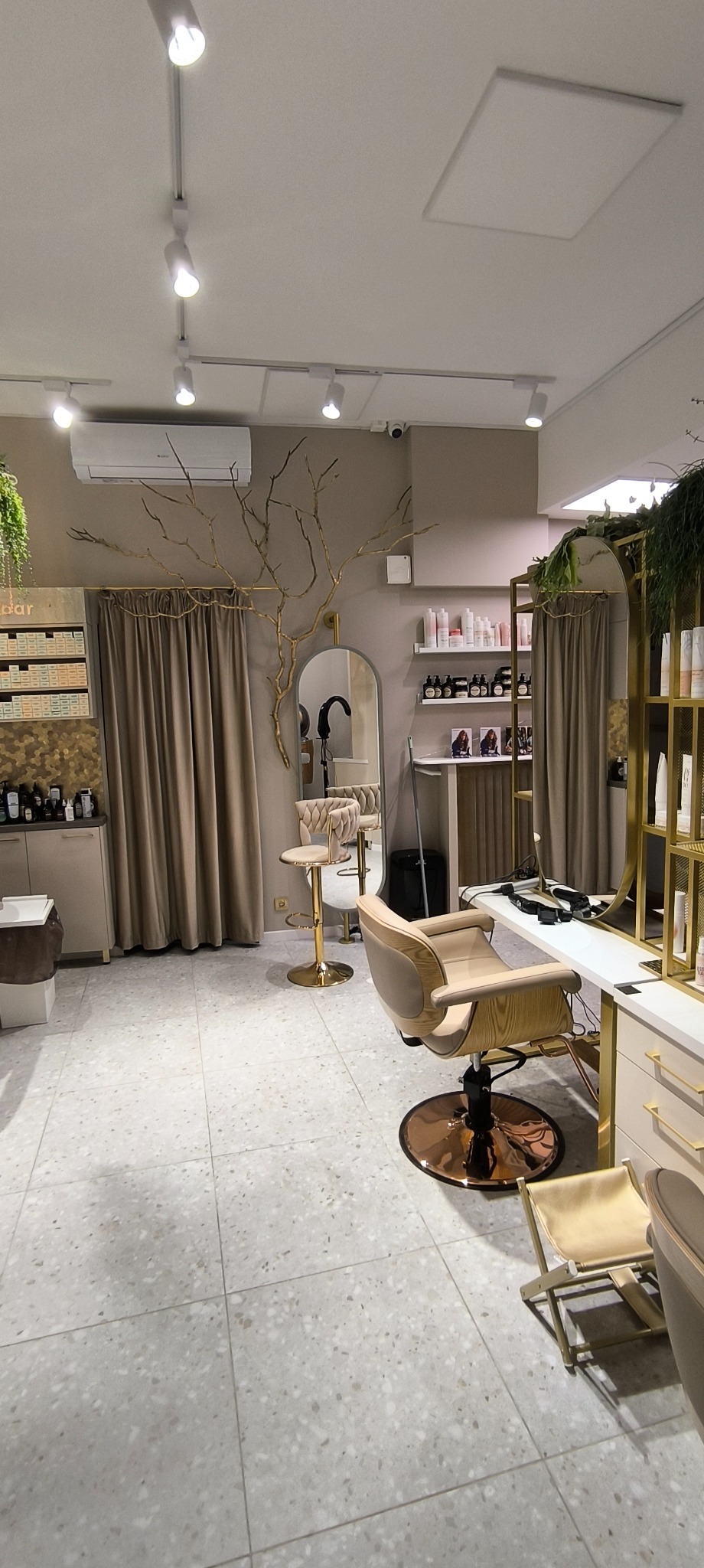 Wnętrze nowoczesnego salonu fryzjerskiego z beżowymi fotelami, złotymi akcentami, lustrami i dekoracyjnym drzewem na ścianie. Widoczne oświetlenie punktowe i jasna podłoga z terazzo.