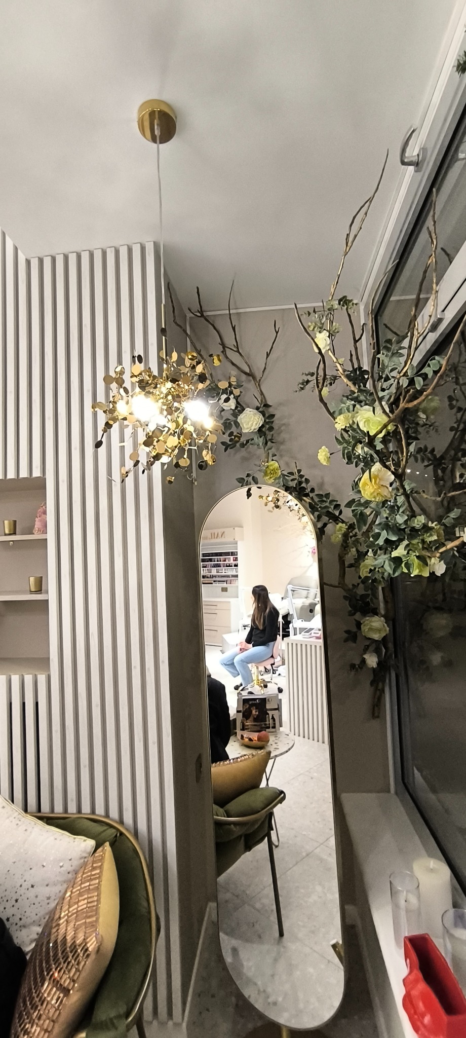 Odbicie w lustrze salonu kosmetycznego z klientką siedzącą na krześle podczas zabiegu manicure, widoczne dekoracje kwiatowe i lampa wisząca.