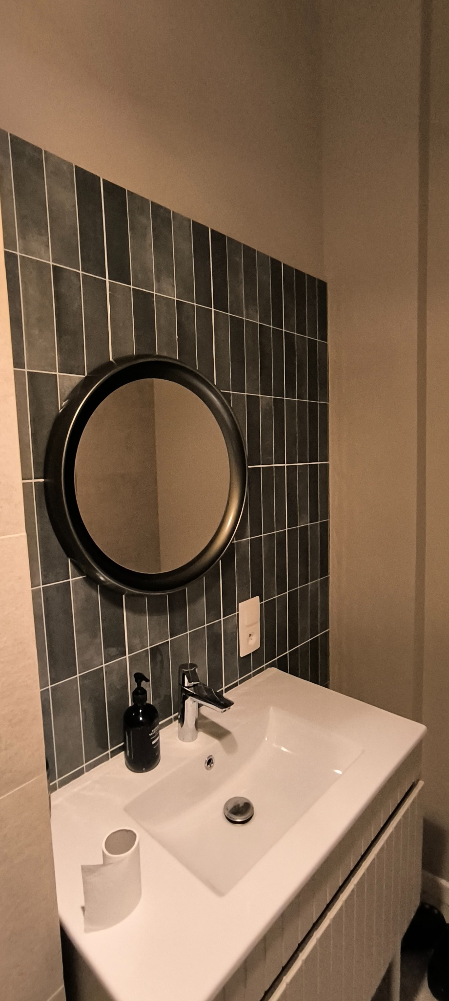 Umywalka z baterią i okrągłym lustrem na tle ściany wyłożonej ciemnymi, pionowymi płytkami z jasną fugą, obok dozownik mydła i rolka papieru toaletowego.