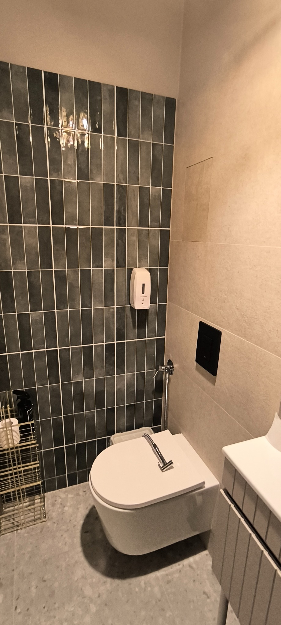 Nowoczesna toaleta podwieszana z bidetką, obok umywalka z szafką, ściana wyłożona ciemnymi, pionowymi płytkami, dozownik na mydło, na podłodze lastryko.