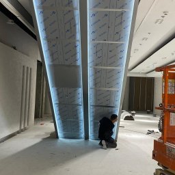 DeLift Łukasz Czechowski - Montaż podświetlanych konstrukcji stalowych w nowoczesnym wnętrzu; pracownik na kolanach przy kolumnie, widoczny podnośnik koszowy.