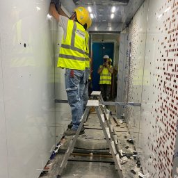 DeLift Łukasz Czechowski - Pracownik w żółtym kasku i kamizelce odblaskowej montuje panele wewnątrz budynku, stojąc na aluminiowej drabinie, widoczne ślady po pracy na podłodze, w tle inny pracownik robi zdjęcie telefonem.