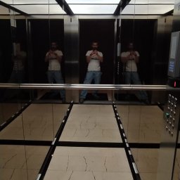 DeLift Łukasz Czechowski - Wnętrze windy z lustrzanymi ścianami, widoczny mężczyzna robiący zdjęcie telefonem, marmurowa podłoga w neutralnych barwach, oświetlenie sufitowe w czarnej ramie.