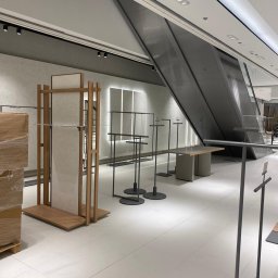 DeLift Łukasz Czechowski - Wnętrze sklepu odzieżowego w trakcie aranżacji: metalowe stojaki na ubrania z okrągłymi podstawami, drewniany regał z jasną płytą, eskalator w tle, minimalistyczny design.